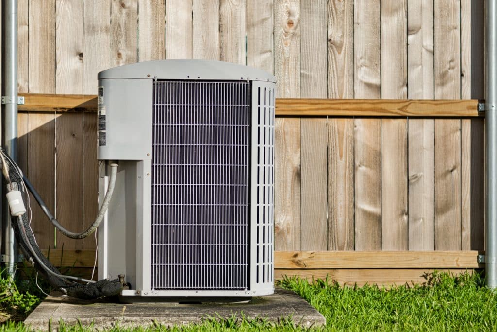Central air conditioning unit in a residential backyard in front of a defocused wooden fence and lawn area with copy space.